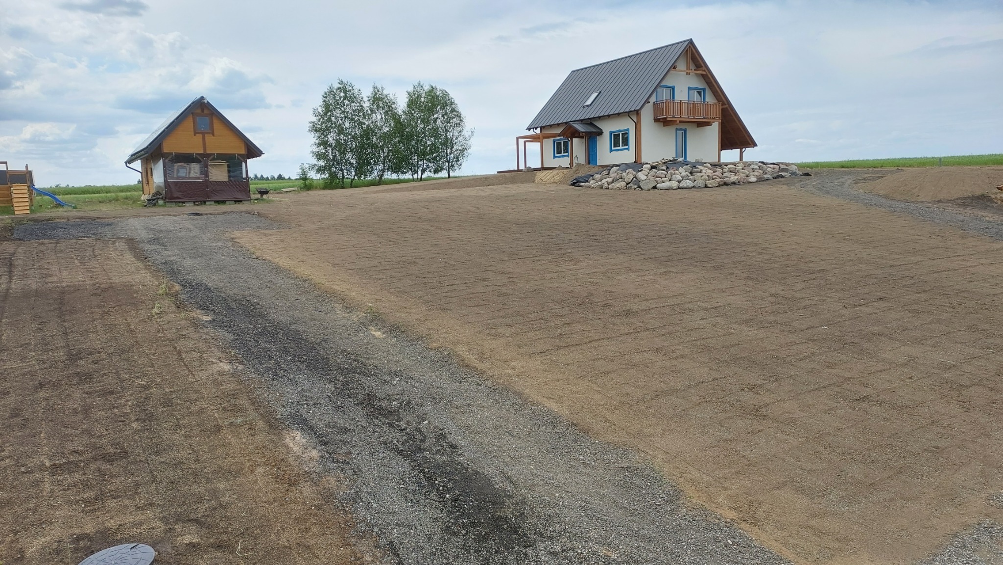 Przygotowany teren pod trawnik z widocznymi śladami po bronowaniu, w tle dom z bali i dom z białego tynku z niebieskimi detalami, kamienie dekoracyjne przy domu.