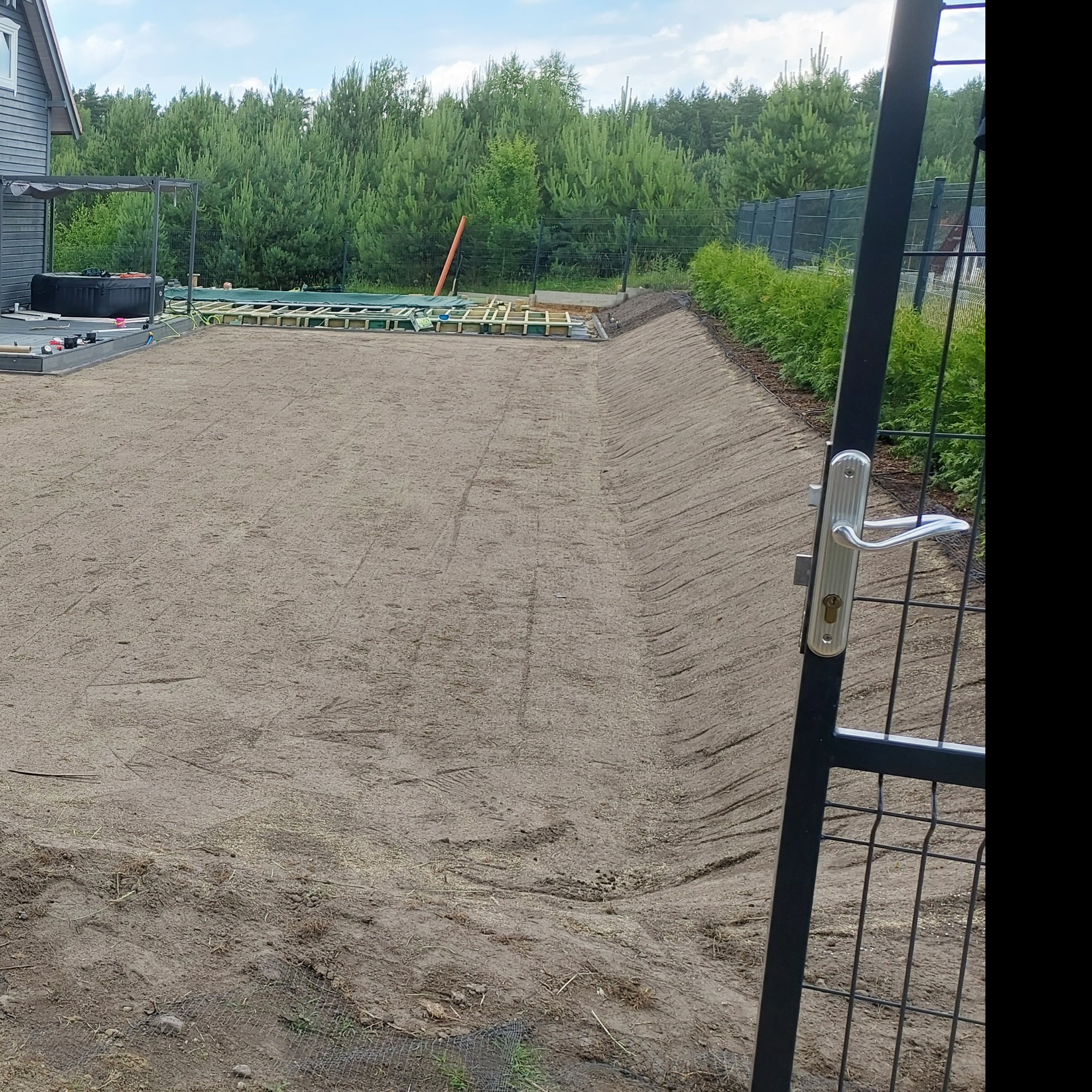 Przygotowany teren pod trawnik z widocznym systemem nawadniania, siatką zabezpieczającą i nowo posadzonymi tujami wzdłuż ogrodzenia.