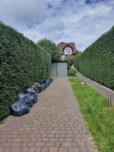 Równy rząd czarnych worków ze ścinkami po strzyżeniu żywopłotu ustawiony wzdłuż ceglanej ścieżki, wysokie żywopłoty po obu stronach, w tle dom z czerwonym dachem i szara blaszana budka.