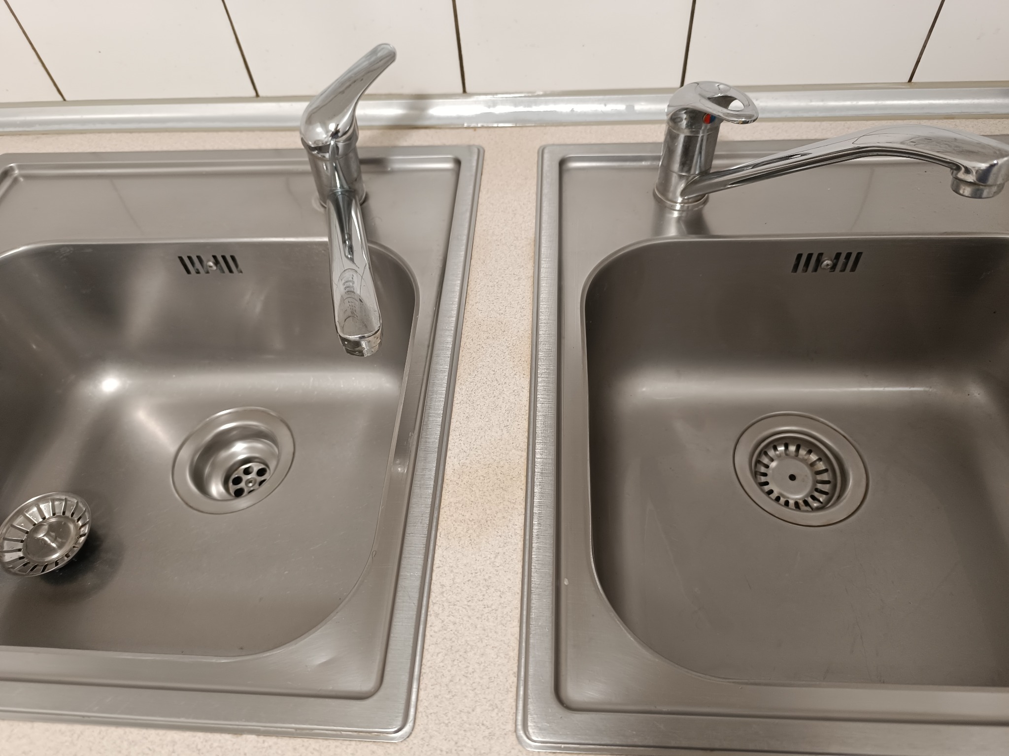 Dwa stalowe zlewozmywaki z bateriami, jeden z sitkiem obok odpływu, na blacie z drobnym wzorem.