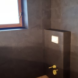 Wojciech Gąsienica - Wykończona łazienka z ciemnymi, marmurkowymi płytkami na ścianach, drewnopodobną podłogą i oknem z drewnianą ramą. Widoczne żółte zawory przy zabudowie WC.