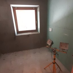 Wojciech Gąsienica - Świeżo otynkowane ściany w pokoju z oknem dachowym, jedna ściana wykończona płytą kartonowo-gipsową, na podłodze leży szpachelka, a pomieszczenie oświetla lampa budowlana na trójnogu.