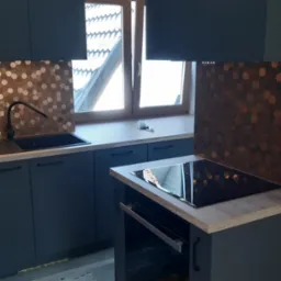 Nowoczesna kuchnia w odcieniach szarości z miedzianą mozaiką heksagonalną na ścianie nad blatem, czarny zlew i bateria, płyta indukcyjna z piekarnikiem, okno z widokiem na dach budynku.