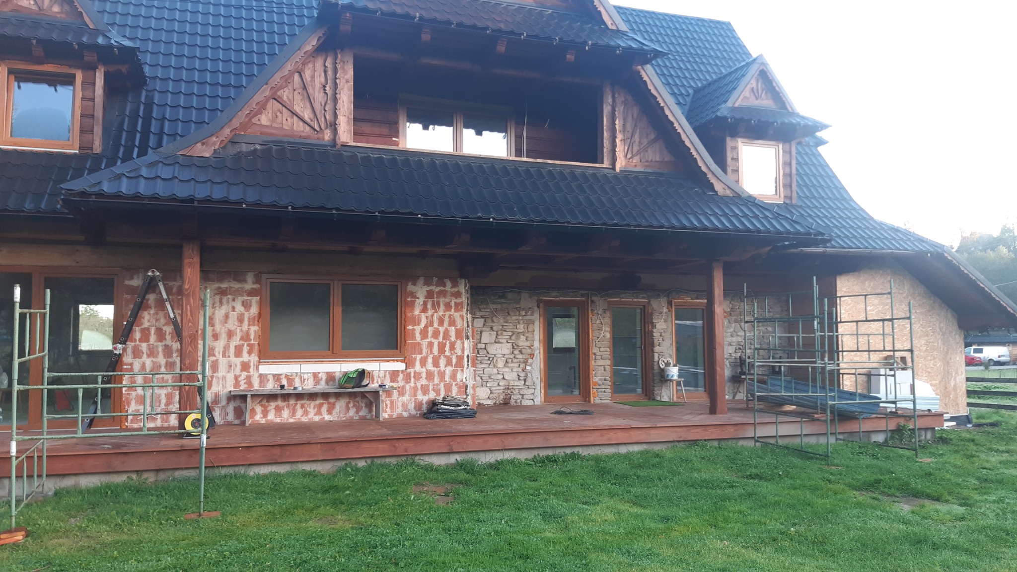 Dom w trakcie prac elewacyjnych: fragment ściany z cegły, fragment z kamienia, drewniana weranda, rusztowania i drabina oparta o ścianę, czarna dachówka.