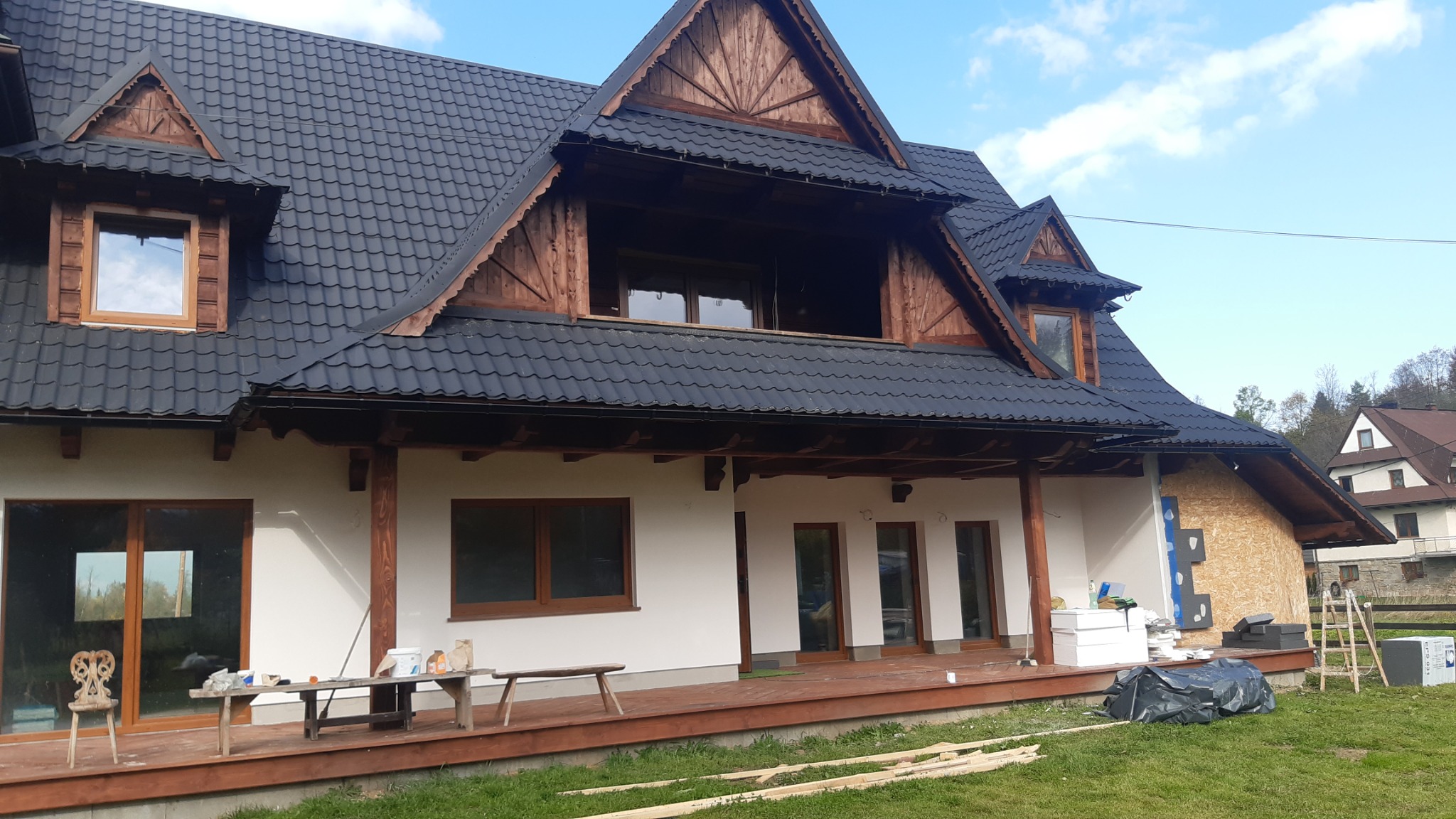 Dom z ciemną dachówką w trakcie prac wykończeniowych elewacji, widoczne drewniane elementy dekoracyjne i rusztowanie z boku budynku.