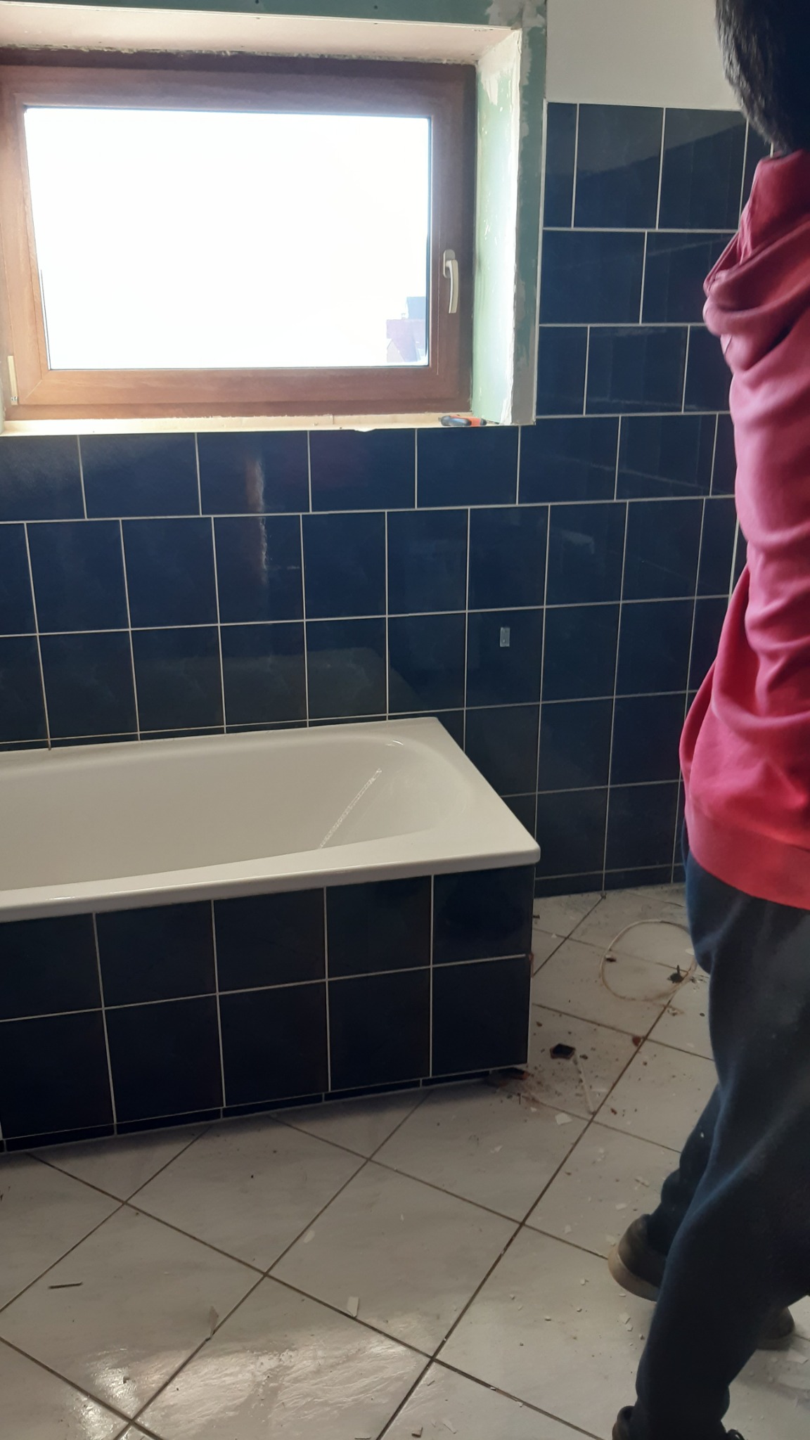Łazienka w trakcie remontu: widok na wannę obłożoną czarnymi płytkami, okno z drewnianą ramą i fragment osoby w różowej bluzie, podłoga częściowo pokryta białymi płytkami w kratkę, widoczne ślady...