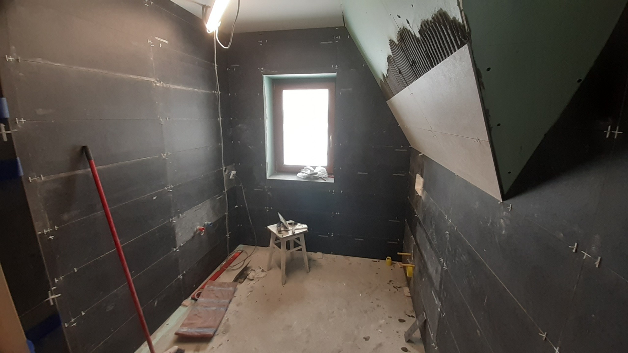 Wnętrze łazienki w trakcie remontu: ciemne płytki ułożone na ścianach z krzyżykami dystansowymi, fragment ściany z płytą karton-gips w górnym rogu, okno z białymi ręcznikami, taboret z laptopem...