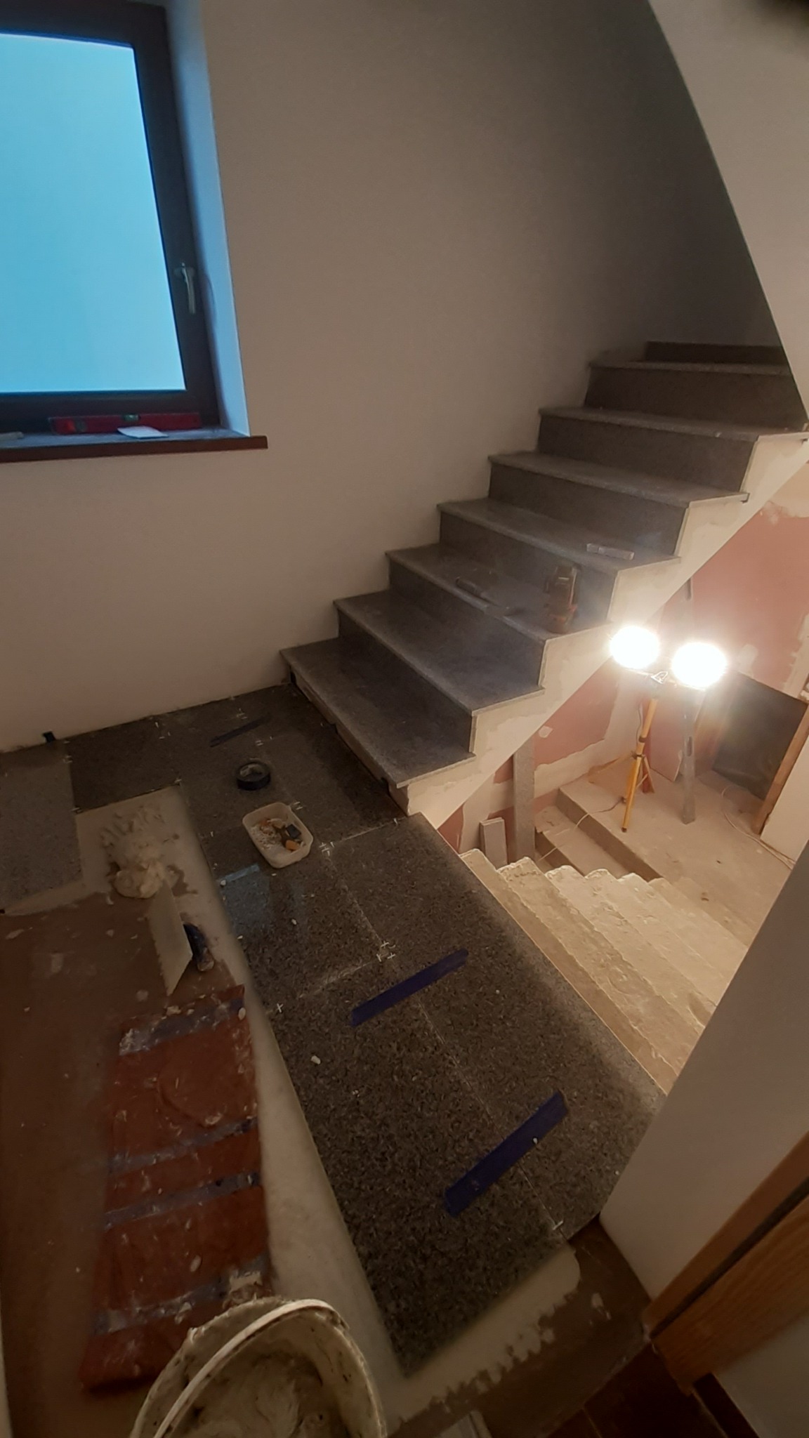 Montaż kamiennych płytek na schodach wewnętrznych, widoczne narzędzia i lampa budowlana, etap prac wykończeniowych.
