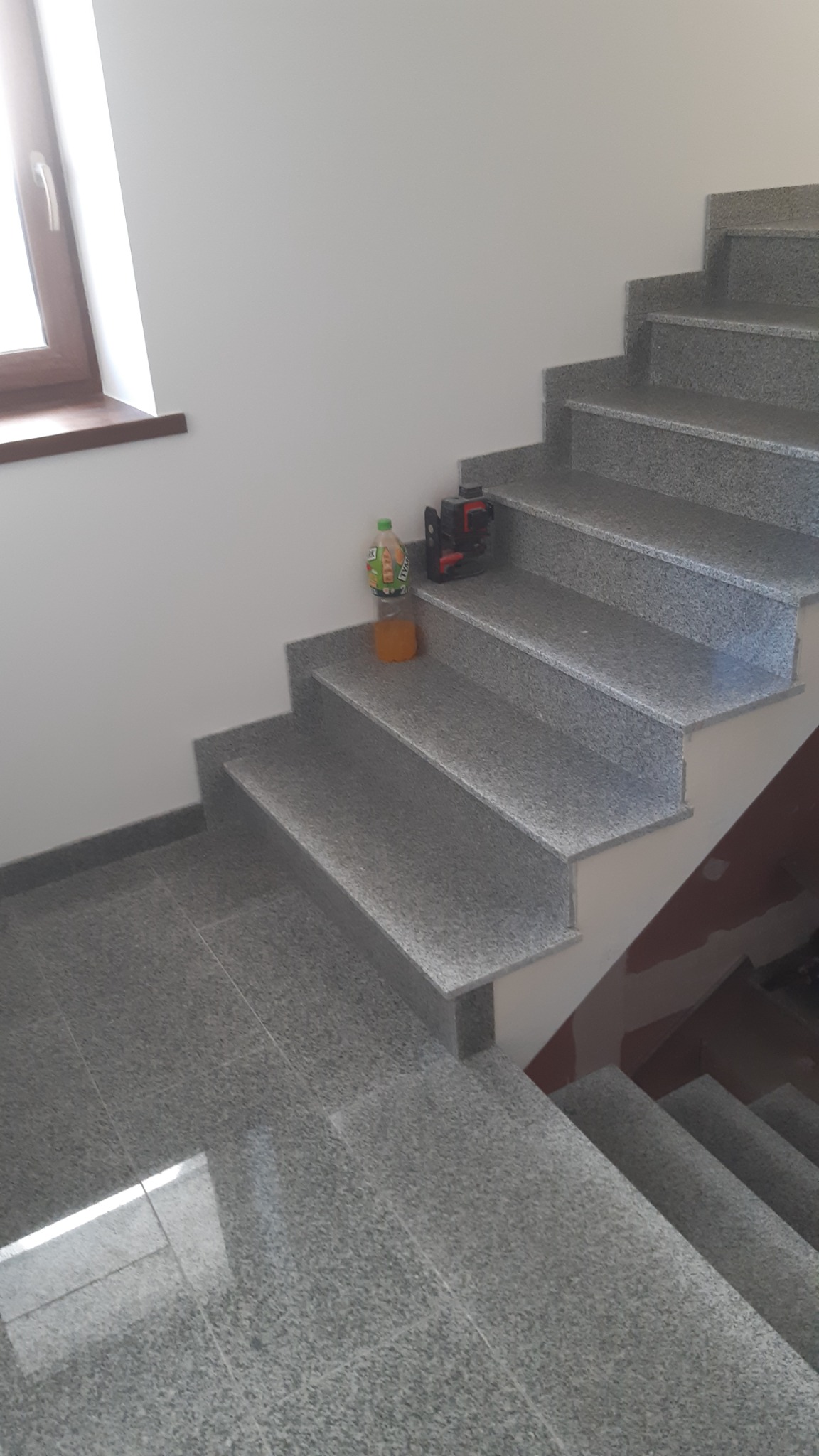 Schody i podłoga wyłożone płytkami imitującymi kamień, z butelką soku i laserowym niwelatorem na stopniu.