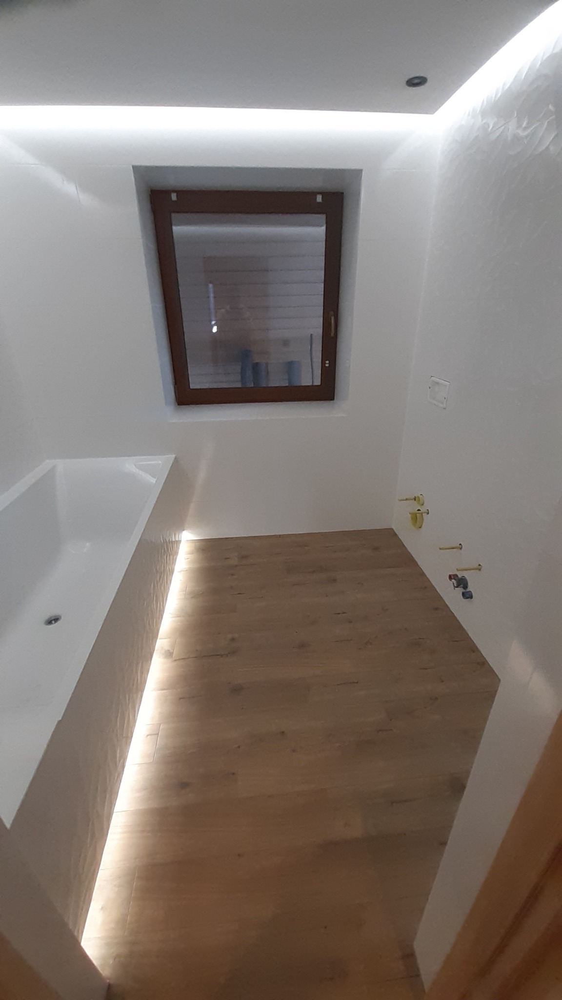 Nowoczesna łazienka w Nowym Targu z drewnopodobną podłogą, białą wanną, oknem w brązowej ramie i subtelnym oświetleniem LED na suficie i wzdłuż wanny. Widoczne przyłącza wodne na ścianie.