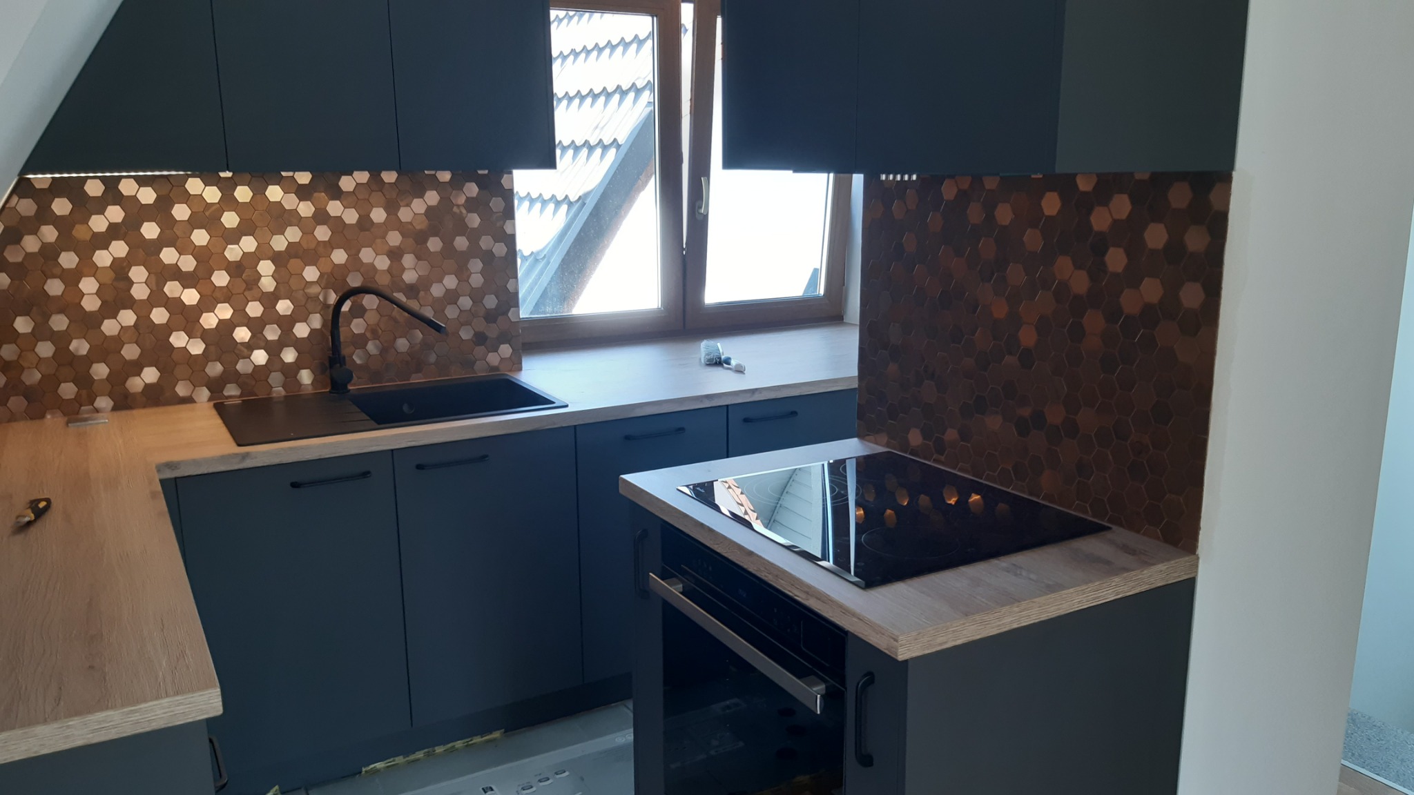 Nowoczesna kuchnia w odcieniach szarości z miedzianą mozaiką heksagonalną na ścianie nad blatem, czarny zlew i bateria, płyta indukcyjna z piekarnikiem, okno z widokiem na dach budynku.