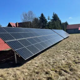 Rząd paneli słonecznych na metalowej konstrukcji ustawiony na trawiastym wzgórzu z widokiem na domy z czerwonymi dachami w tle, błękitne niebo.