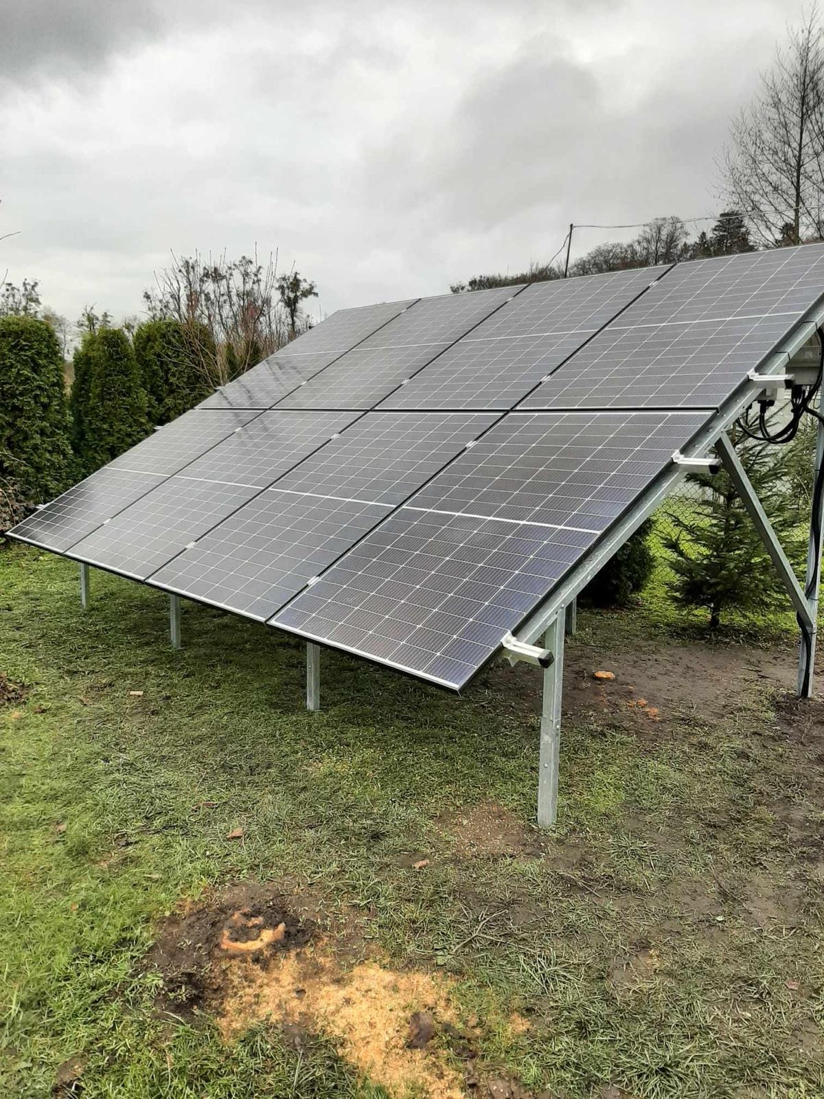 Instalacja fotowoltaiczna na metalowej konstrukcji, ustawiona na trawniku w pochmurny dzień. Widoczne przewody i elementy montażowe.
