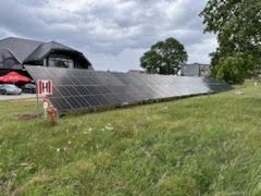 Rząd paneli słonecznych zamontowanych na trawiastym terenie, z budynkiem w tle i pochmurnym niebem. Widoczny słupek z czerwoną skrzynką.