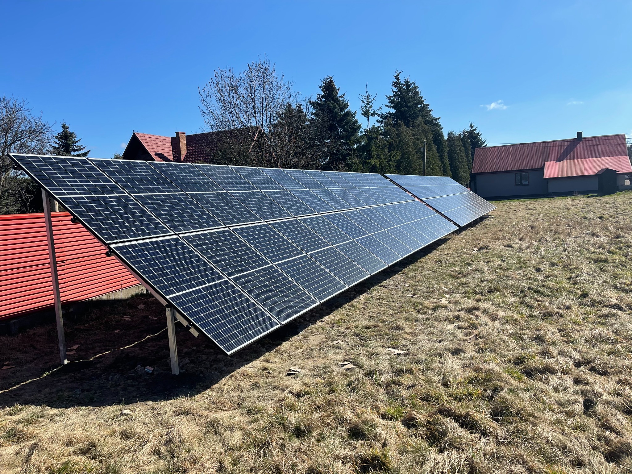 Rząd paneli słonecznych na metalowej konstrukcji ustawiony na trawiastym wzgórzu z widokiem na domy z czerwonymi dachami w tle, błękitne niebo.