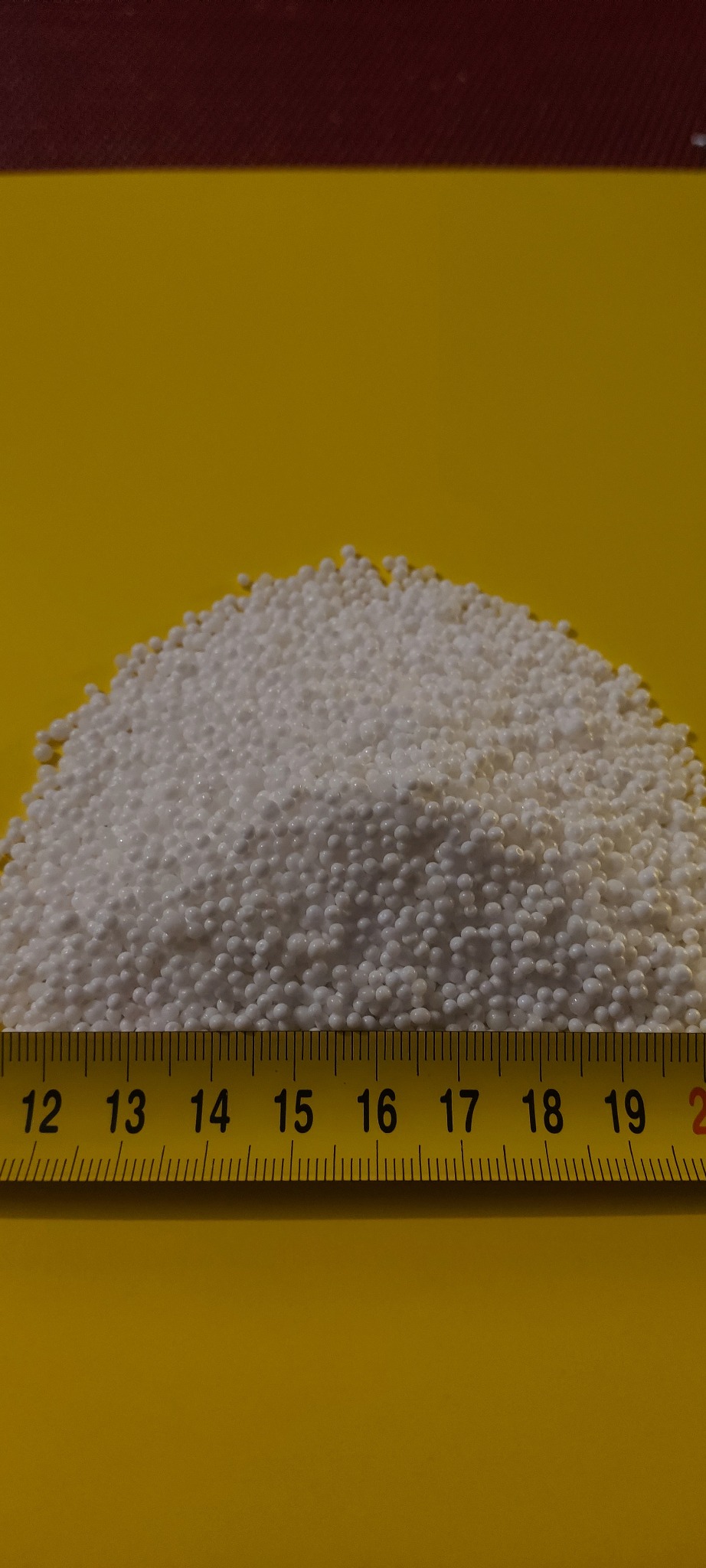 Górka białych granulek, prawdopodobnie paliwa stałego, zmierzona żółtą miarką od 12 do 20 centymetrów na żółtym tle.