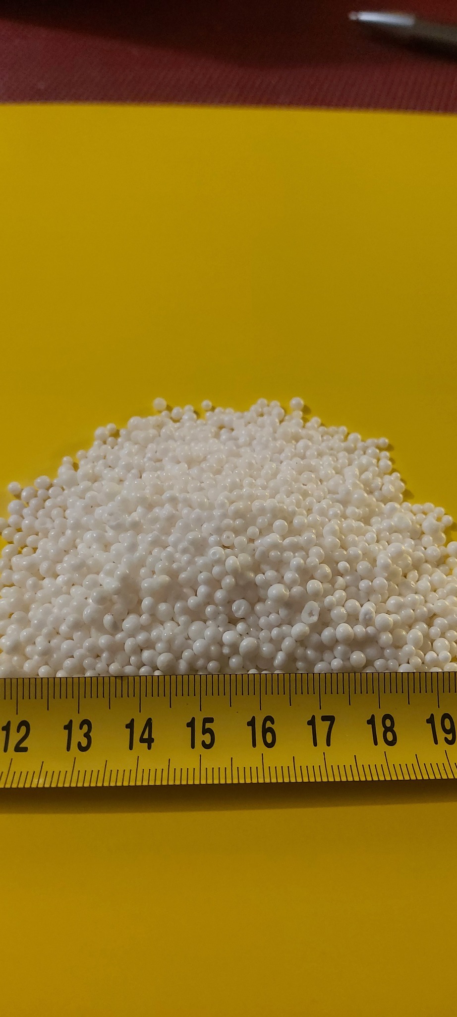 Białe kulki ekogroszku na żółtym tle, zmierzone żółtą miarką od 12 do 19 centymetrów.