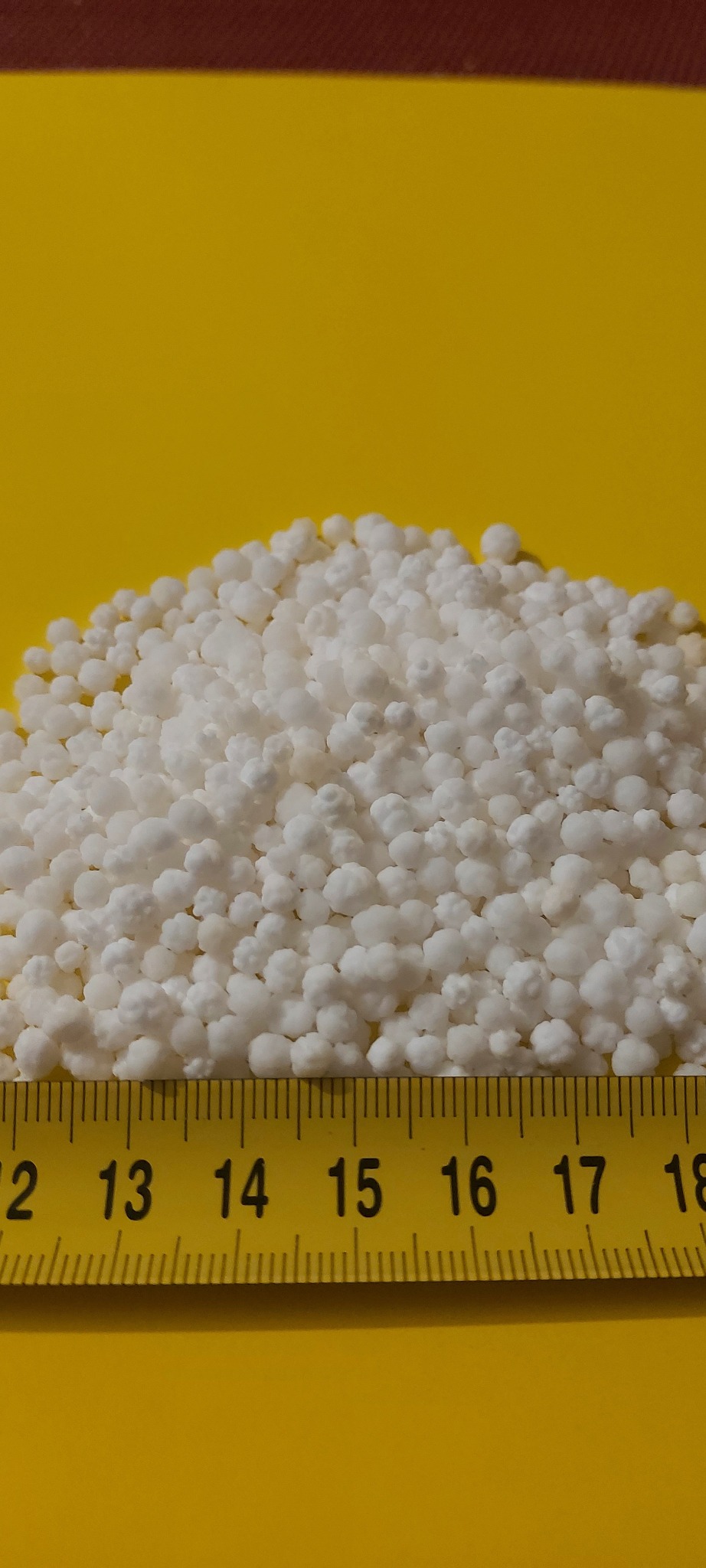 Białe granulki paliwa stałego ułożone na żółtym tle, z widoczną miarką w centymetrach, wskazującą rozmiar granulek.