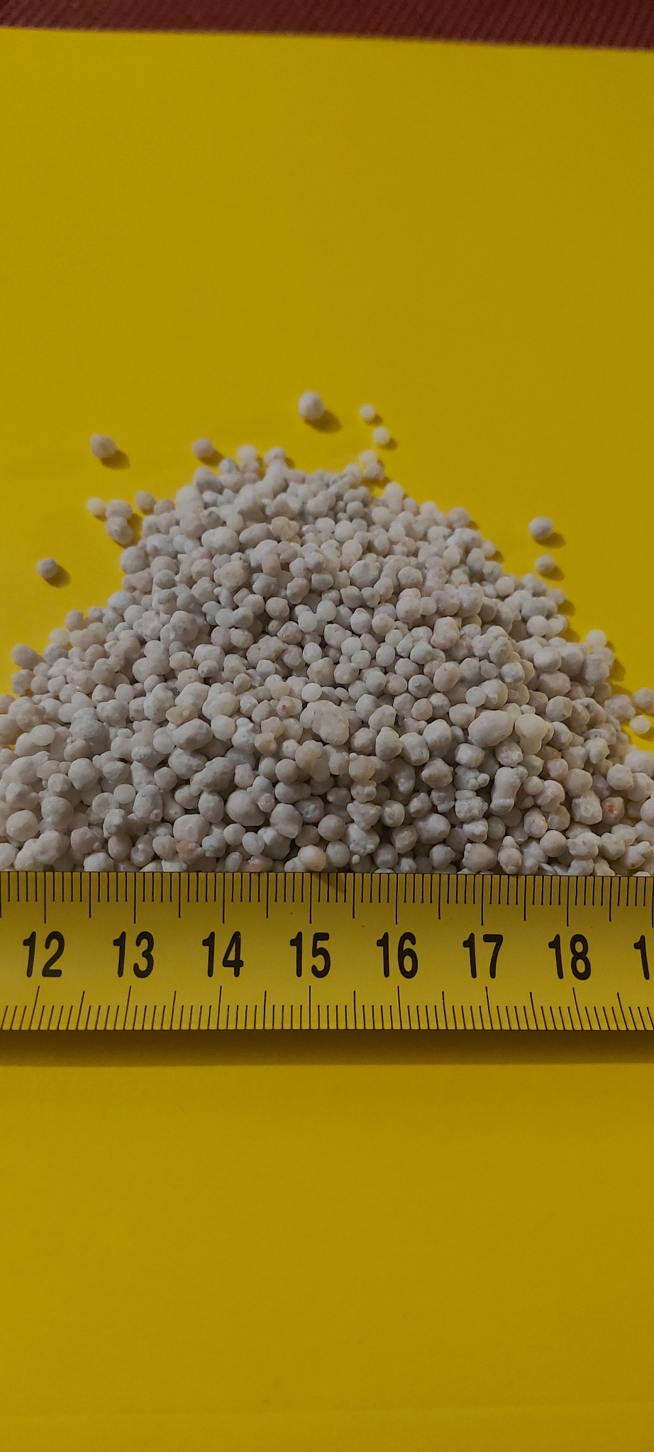 Górka białoszarego granulatu, zmierzona żółtą miarką od 12 do 19 cm na jaskrawożółtym tle.