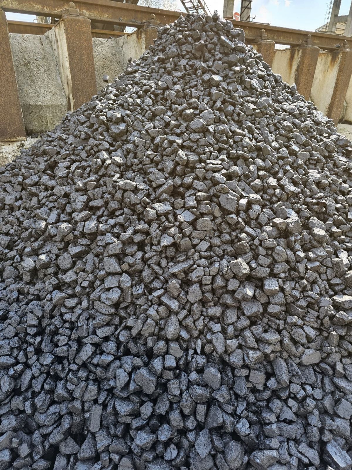 Kopuła błyszczącego, czarnego paliwa stałego na tle betonowych konstrukcji i stalowych belek pod zadaszeniem.