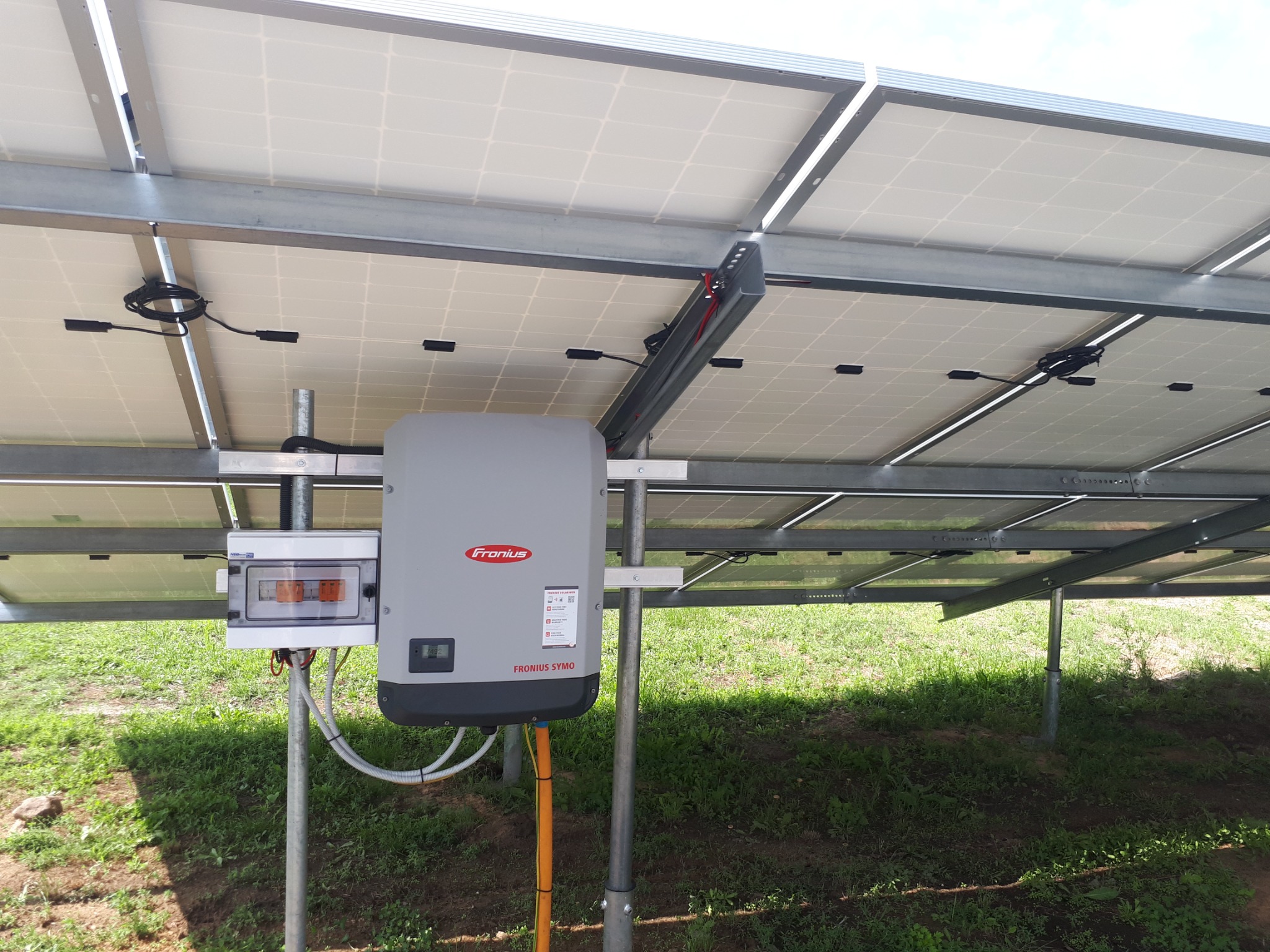 Widok od spodu na zainstalowane panele fotowoltaiczne z widocznym inwerterem Fronius Symo i skrzynką zabezpieczeń na metalowej konstrukcji, na tle trawy.