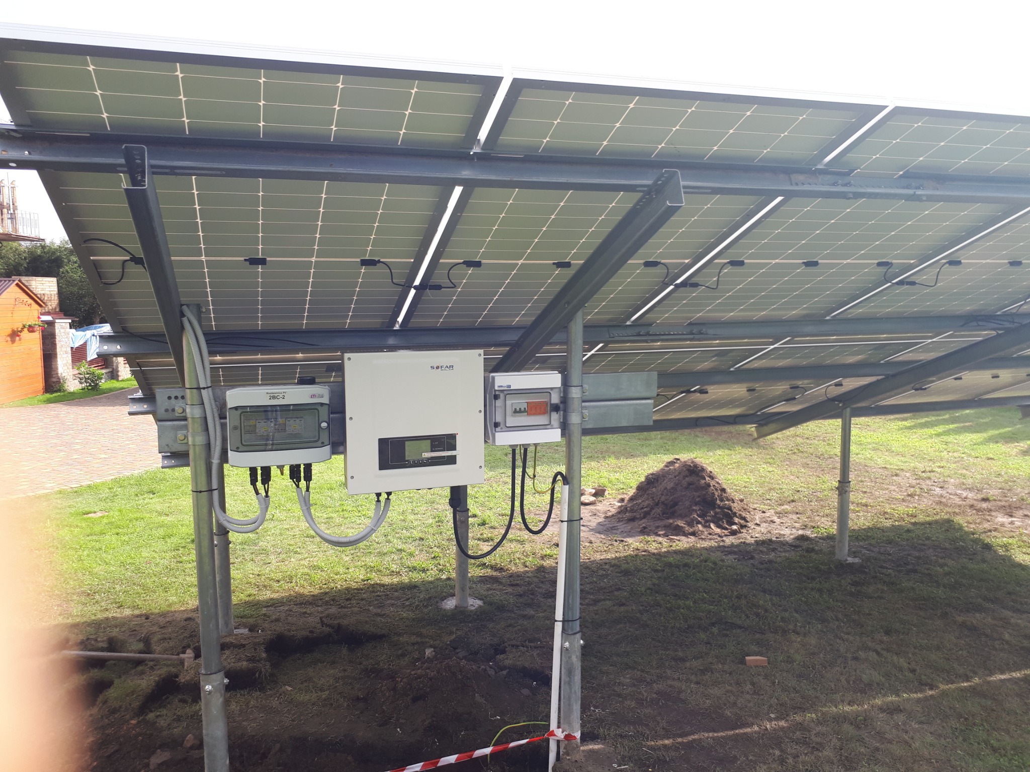 Widok od spodu na panele fotowoltaiczne zamontowane na metalowej konstrukcji, z widocznymi elementami instalacji elektrycznej, takimi jak inwerter i skrzynki zabezpieczeń, na tle trawnika z kopcem...