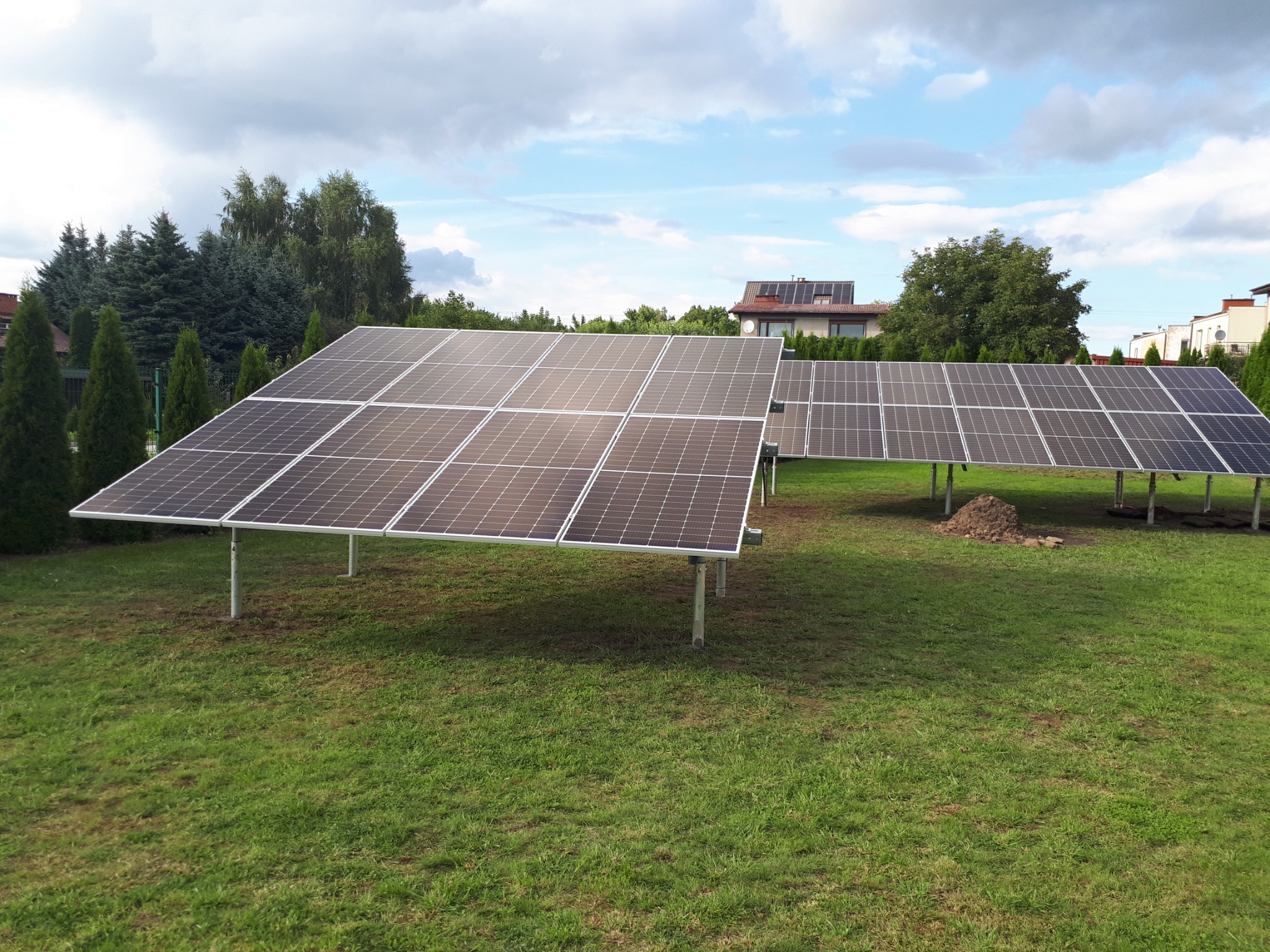 Zestaw paneli słonecznych na trawniku w ogrodzie, widoczne domy w tle, pochmurne niebo.