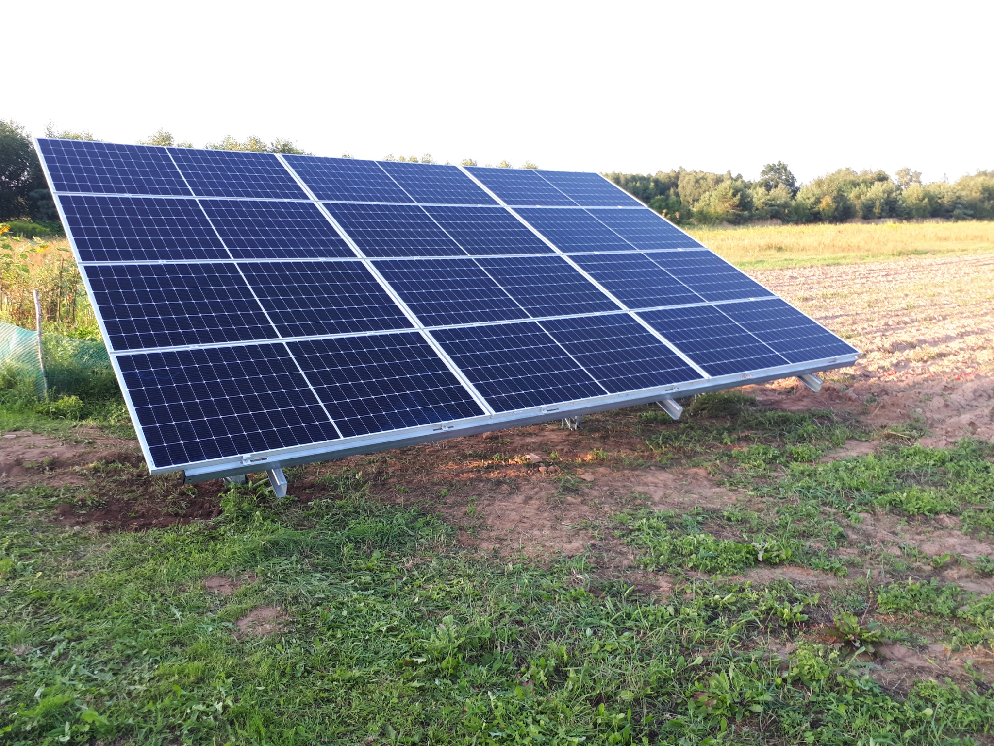 Panel słoneczny z widocznymi mocowaniami na trawiastym terenie z polną roślinnością w tle.
