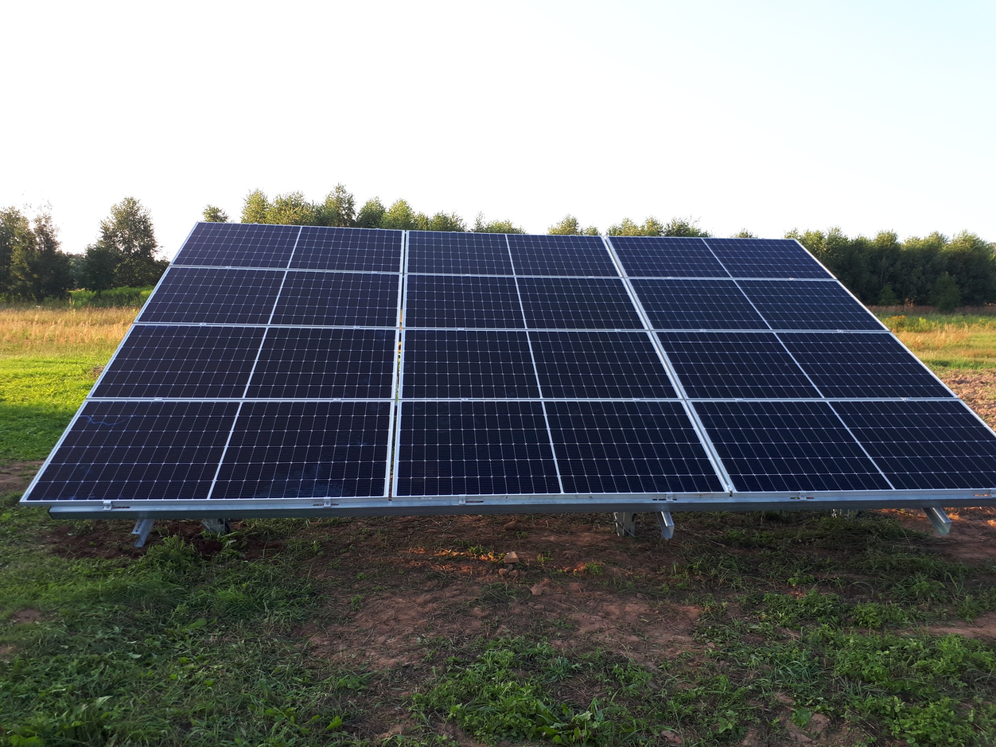 Panel słoneczny z czarnymi ogniwami i srebrną ramą, zamontowany na metalowej konstrukcji na trawiastym terenie z widokiem na drzewa w tle.