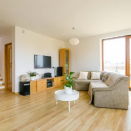 Jasne wnętrze salonu z drewnianymi schodami, parkietem, narożną kanapą i dużym oknem tarasowym z widokiem na ogród.