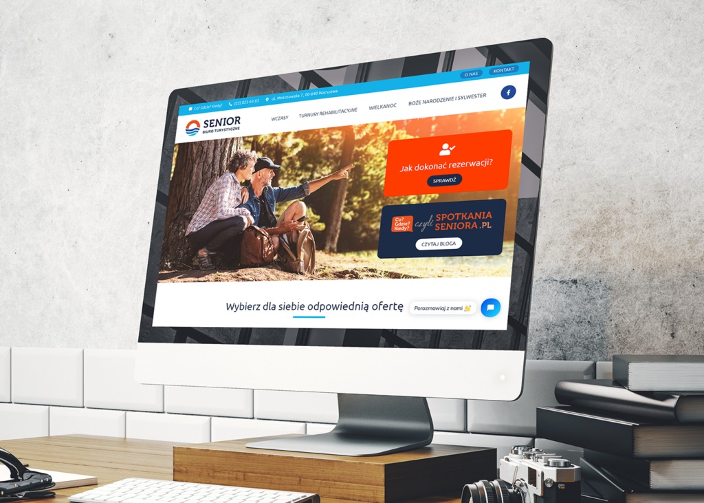 Monitor komputera wyświetlający stronę internetową biura turystycznego Senior z ofertami wczasów i turnusów rehabilitacyjnych dla seniorów, na biurku z książkami i aparatem.