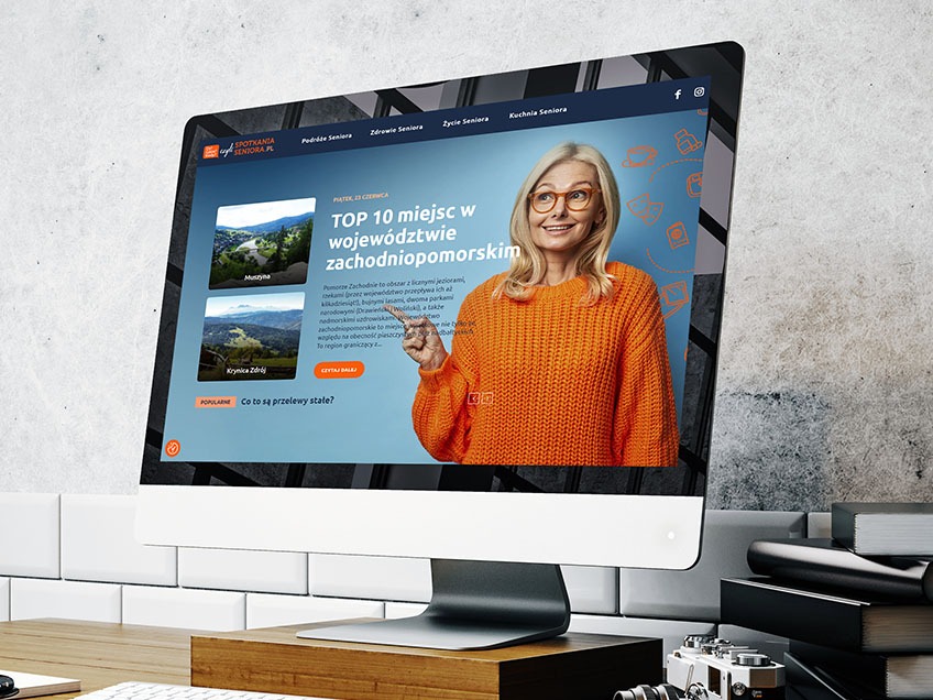 Monitor iMac wyświetlający stronę internetową spotkaniaseniora.pl z artykułem o atrakcjach turystycznych województwa zachodniopomorskiego, obok monitora leżą książki i aparat fotograficzny.