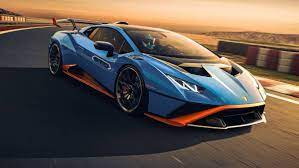 Błękitny samochód sportowy Lamborghini Huracan STO z pomarańczowymi akcentami, pędzący po torze wyścigowym podczas zachodu słońca, z widocznymi elementami zabezpieczeń toru po prawej stronie.