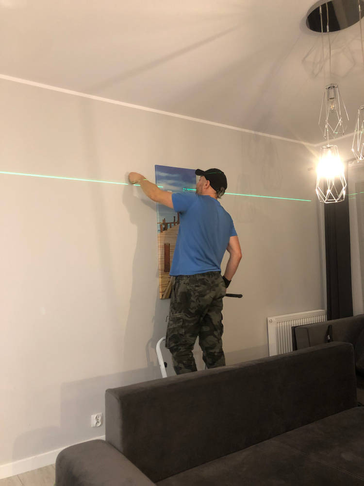 Mężczyzna w czapce i spodniach moro, stojący na drabince, wyznacza poziom laserem na ścianie podczas wieszania obrazu w salonie. Widoczny fragment sofy i nowoczesna lampa.