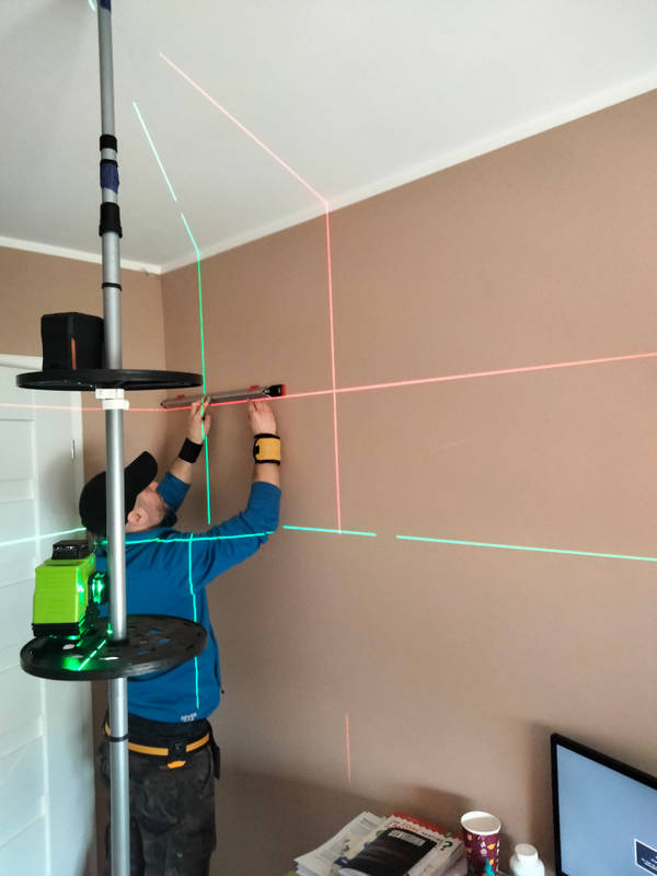 Mężczyzna w bluzie z długim rękawem używa poziomicy laserowej emitującej zielone i czerwone linie na ścianie w pokoju, z widocznym statywem i poziomnicą w ręku.