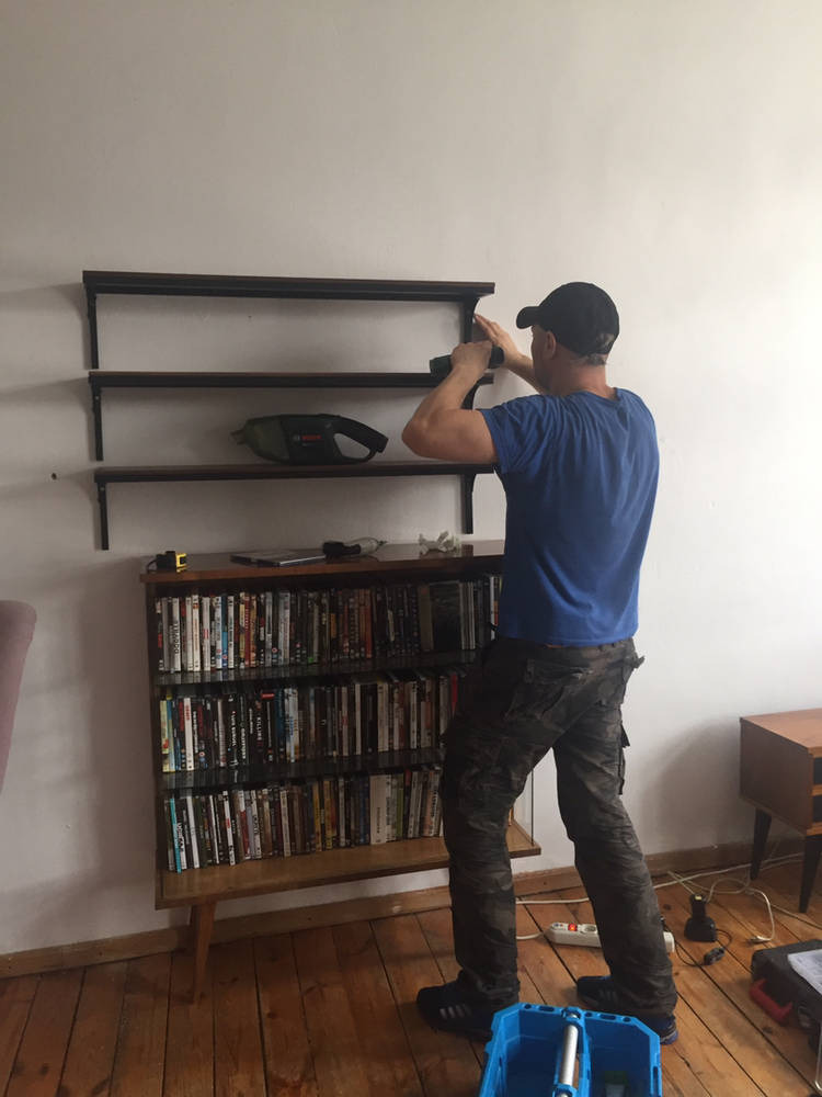 Mężczyzna w czapce i niebieskiej koszulce montuje półki na ścianie w pokoju, używając wiertarki; pod spodem stoi regał z płytami DVD.