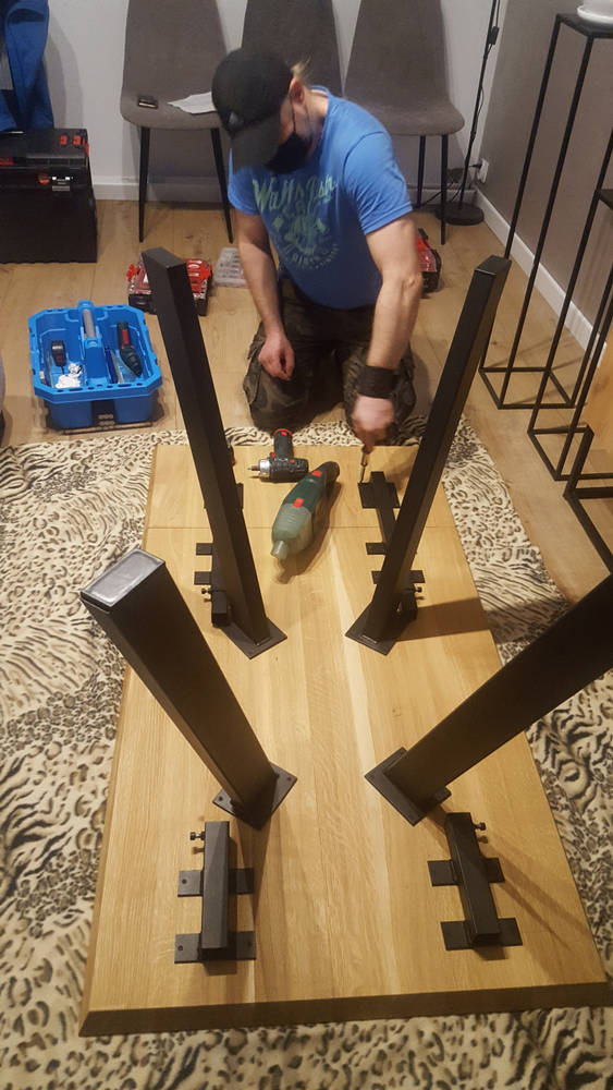 Mężczyzna w maseczce przykręca metalowe nogi do drewnianego blatu stołu za pomocą wkrętarki; widoczny niebieski pojemnik z narzędziami, podłoga pokryta dywanem w panterkę.