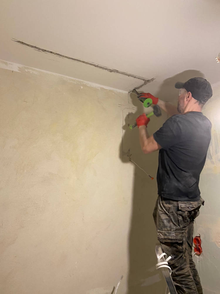 Mężczyzna w czerwonych rękawicach, stojący na drabinie, wykuwa bruzdę w suficie za pomocą dłuta i młotka z zielonym uchwytem, przygotowując miejsce na instalację elektryczną. Widoczne puszki...