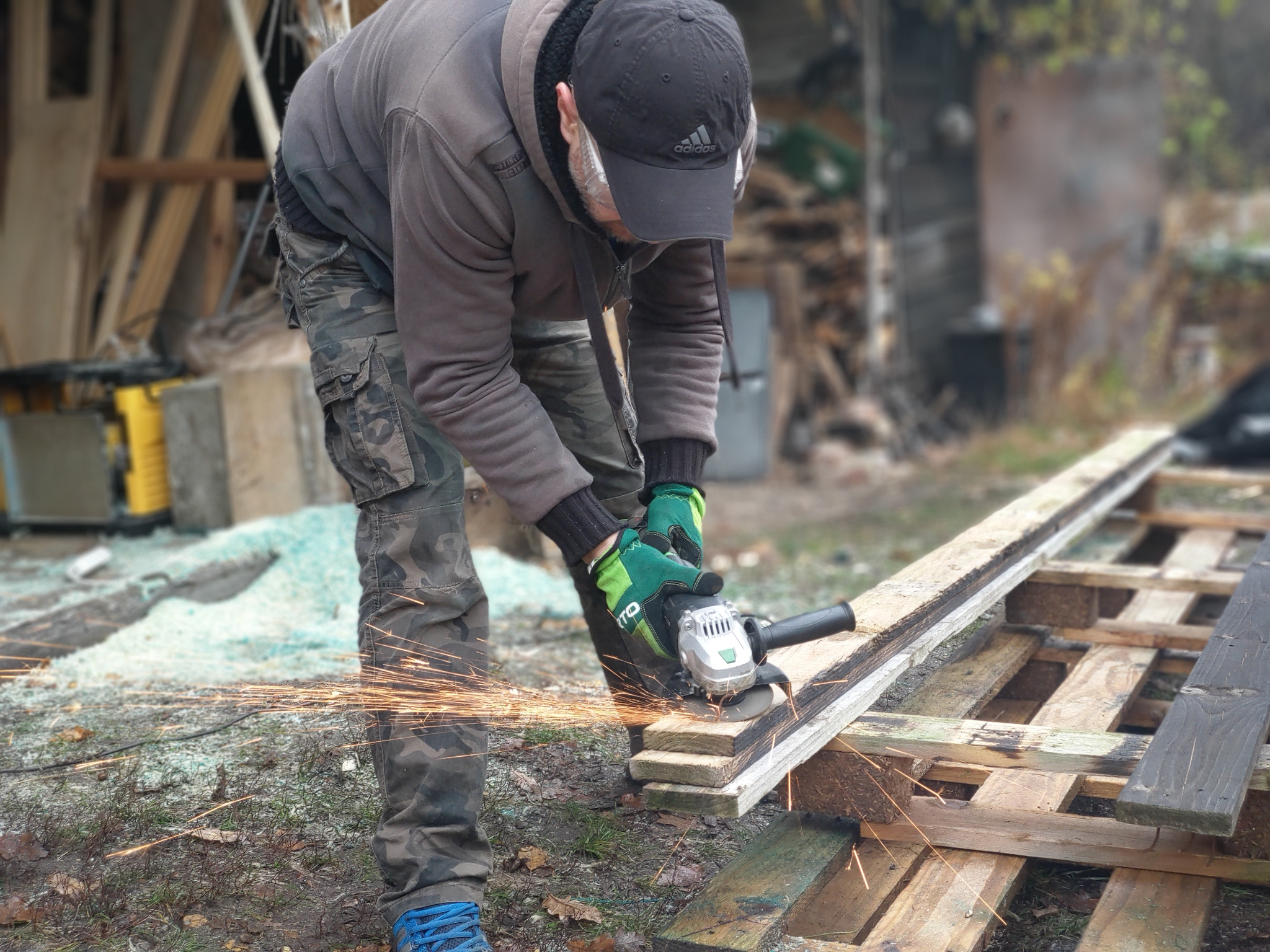 Osoba w czapce Adidas i rękawicach roboczych tnie deskę szlifierką kątową, iskry lecą na boki, w tle widoczne drewniane elementy konstrukcyjne.