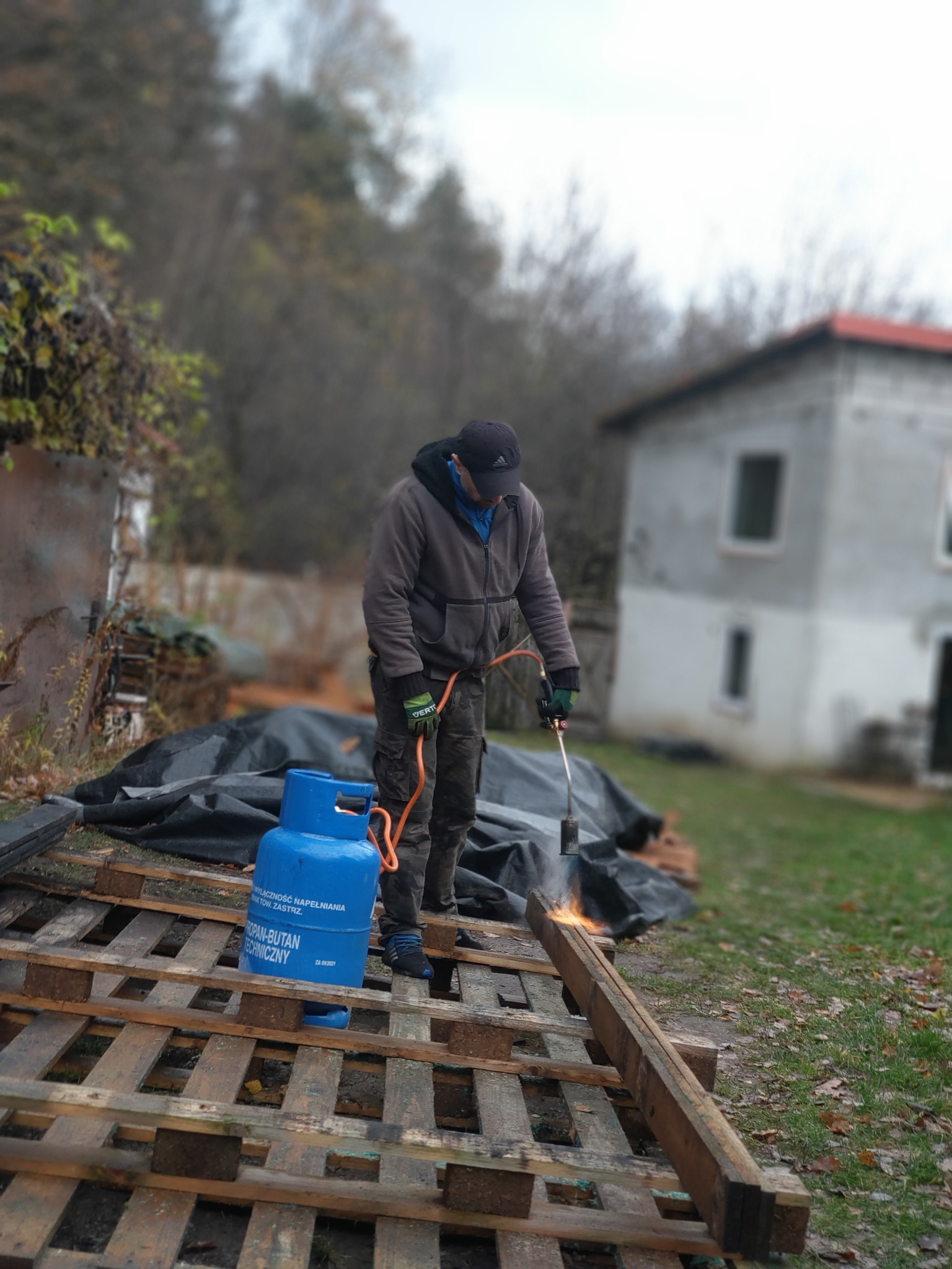 Pracownik w ogrodzie opala drewno z palet za pomocą palnika na gaz propan-butan, przygotowując je do dalszej obróbki. W tle widoczny dom z czerwonym dachem i zieleń.