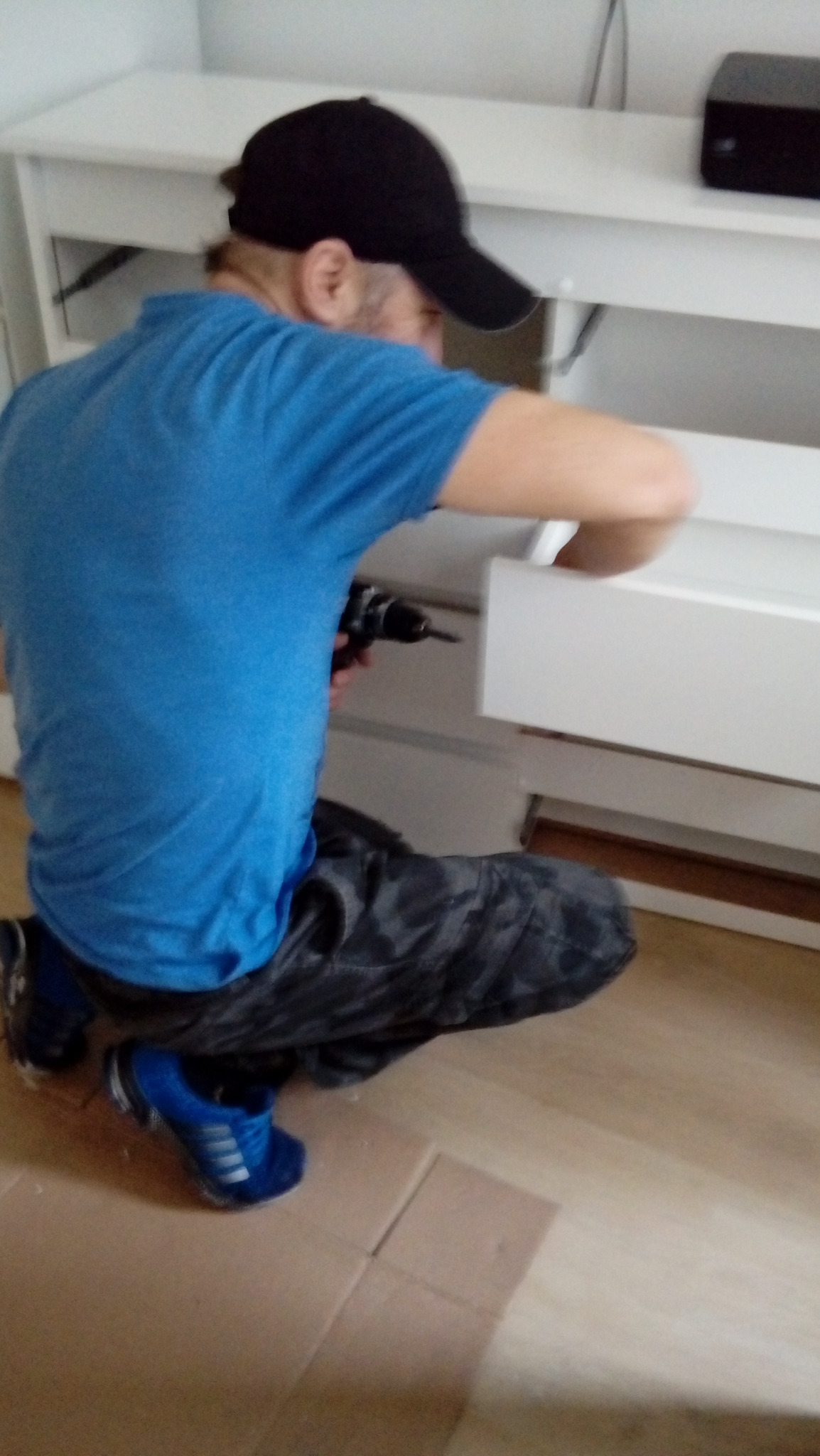 Mężczyzna w niebieskiej koszulce i czapce, na klęczkach, montuje białą szafkę za pomocą wiertarki elektrycznej, na podłodze leżą kartony.