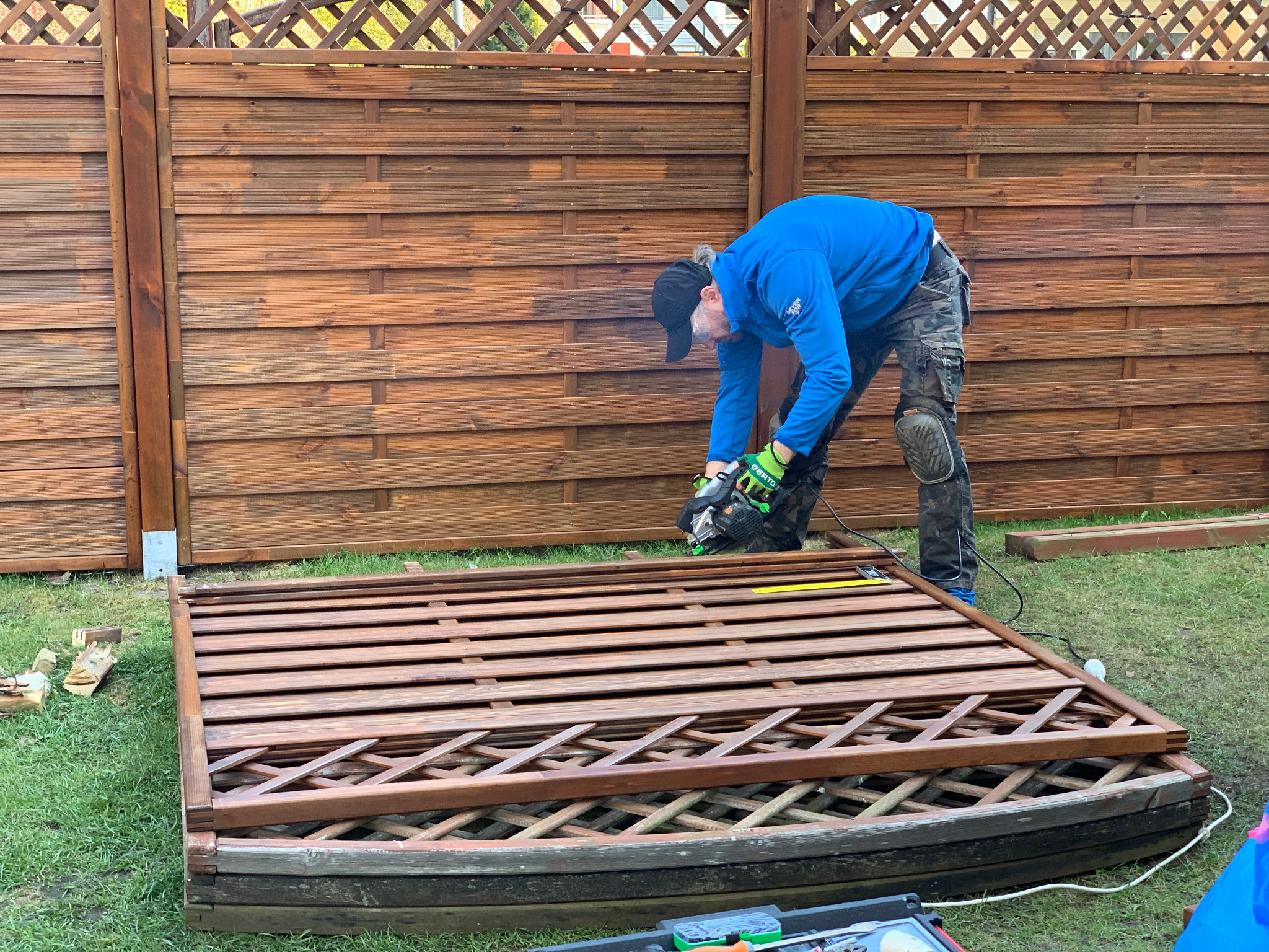 Pracownik w Gdańsku przycina drewnianą kratę ogrodową piłą elektryczną na tle brązowego płotu z desek.