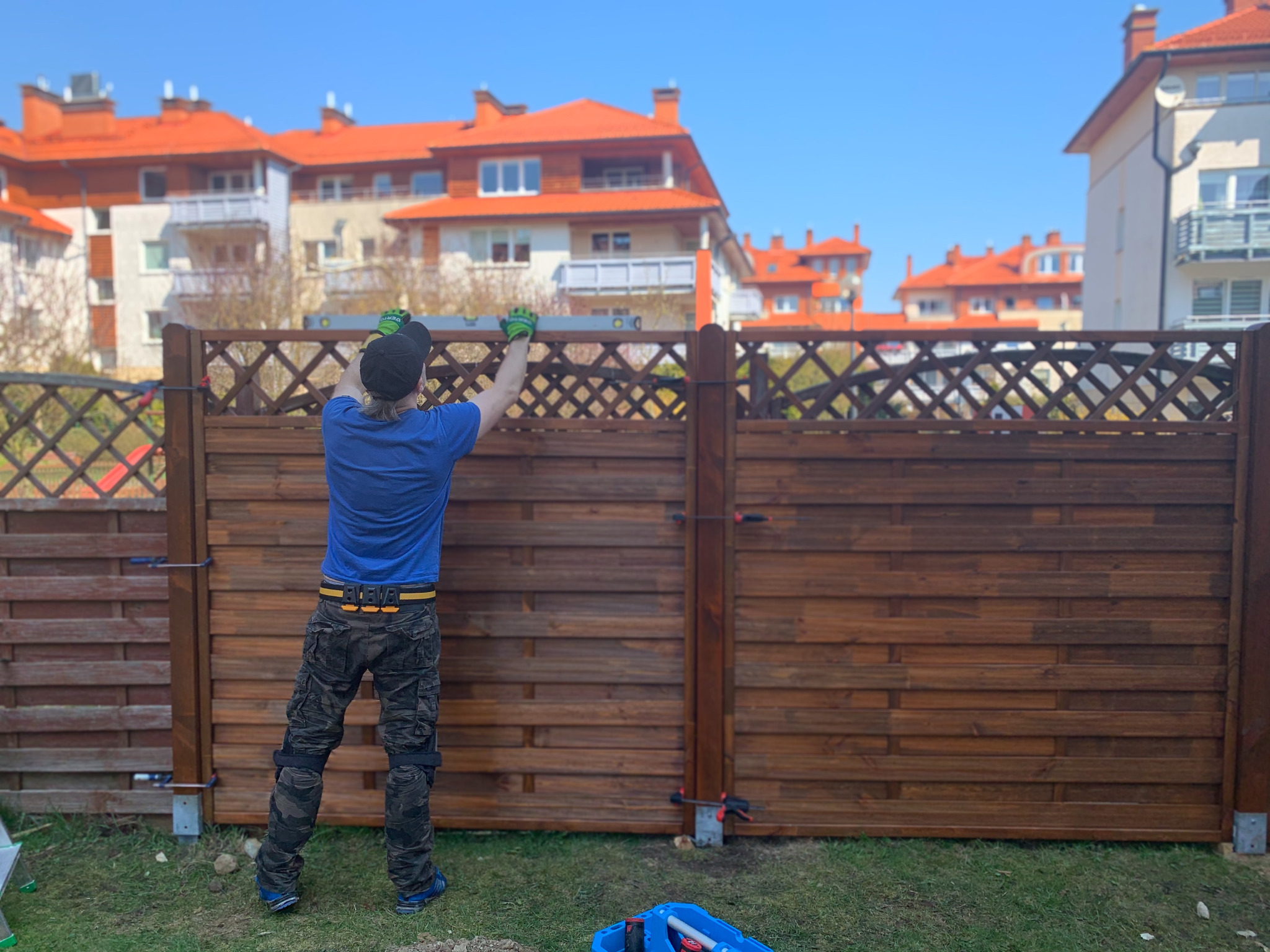 Mężczyzna w czapce i rękawicach roboczych, z poziomicą w rękach, sprawdza poziom nowo postawionego, drewnianego płotu z ozdobną kratką na tle budynków z czerwonymi dachami.