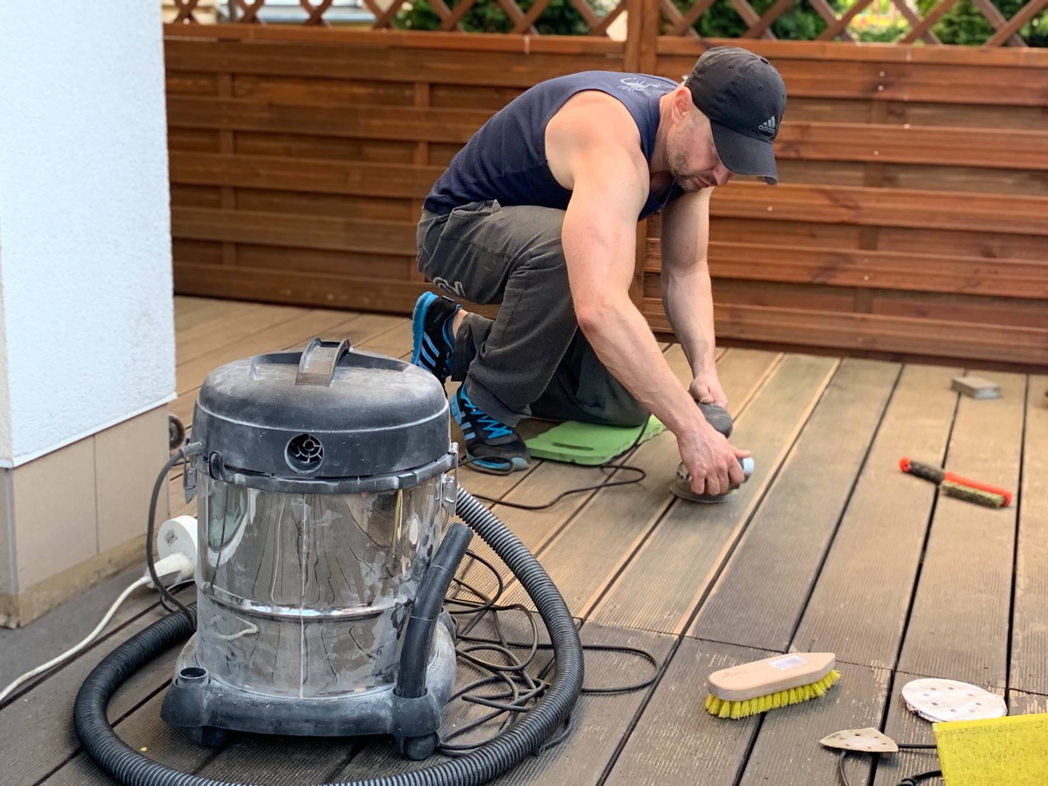 Mężczyzna szlifuje drewnianą podłogę na zewnątrz za pomocą szlifierki oscylacyjnej, obok stoi odkurzacz przemysłowy, a w tle widoczny drewniany płot.