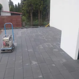 Ułożona szara kostka betonowa na posesji z ubijarką w rogu, widoczne drzewa w tle i fragment budynku.