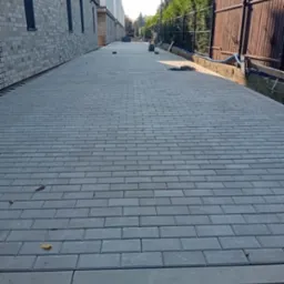 Ułożona szara nawierzchnia z prostokątnej kostki, widok wzdłuż ścieżki, obok budynku z cegły i drewnianego płotu.