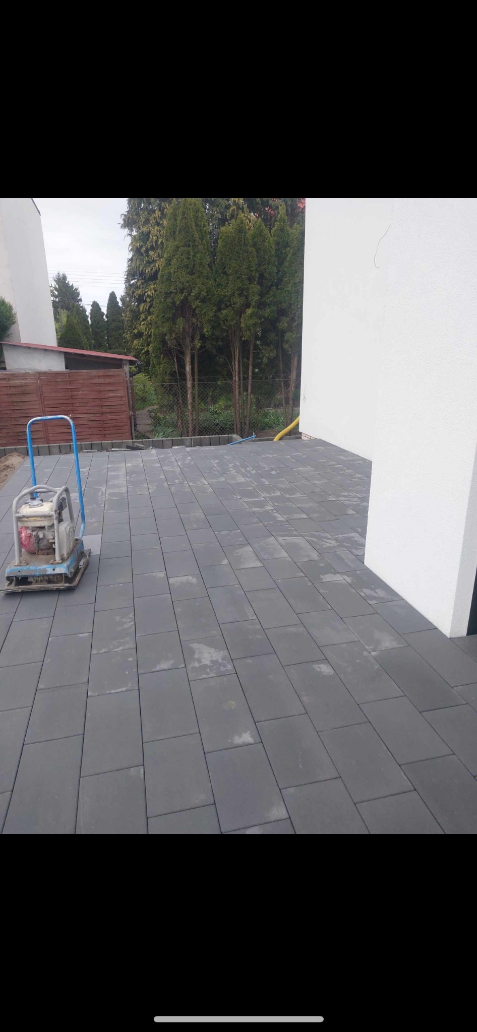 Ułożona szara kostka betonowa na posesji z ubijarką w rogu, widoczne drzewa w tle i fragment budynku.