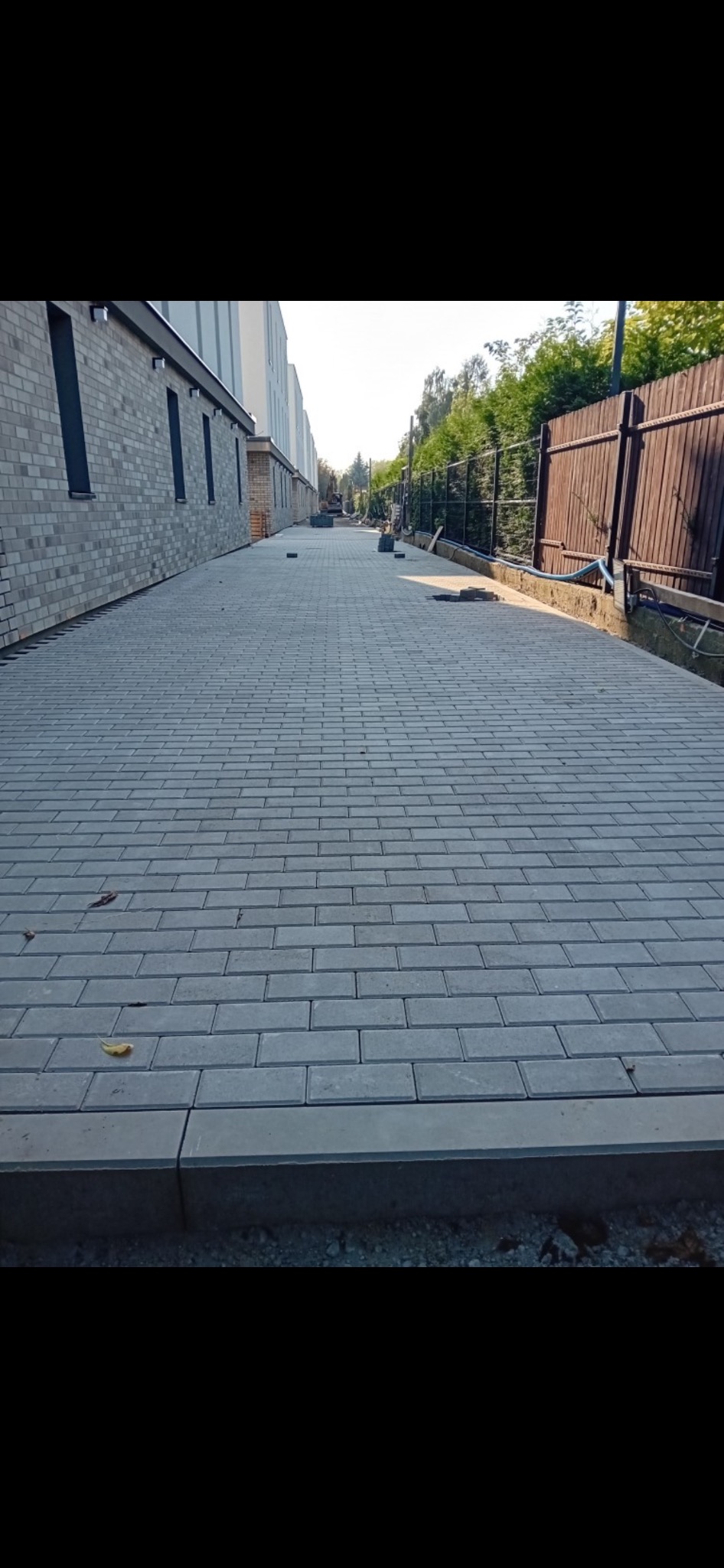 Ułożona szara nawierzchnia z prostokątnej kostki, widok wzdłuż ścieżki, obok budynku z cegły i drewnianego płotu.