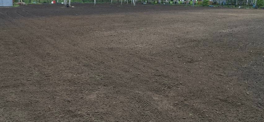 Równy, przygotowany teren pod przyszłą inwestycję, z widocznymi śladami opon i butów, otoczony drzewami.