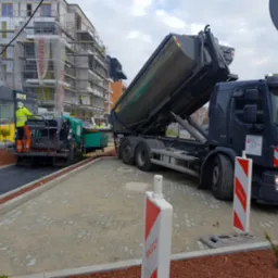 Rozładunek asfaltu z wywrotki do rozściełacza podczas układania nowej nawierzchni chodnika z kostki brukowej, w tle budynek w rusztowaniach.